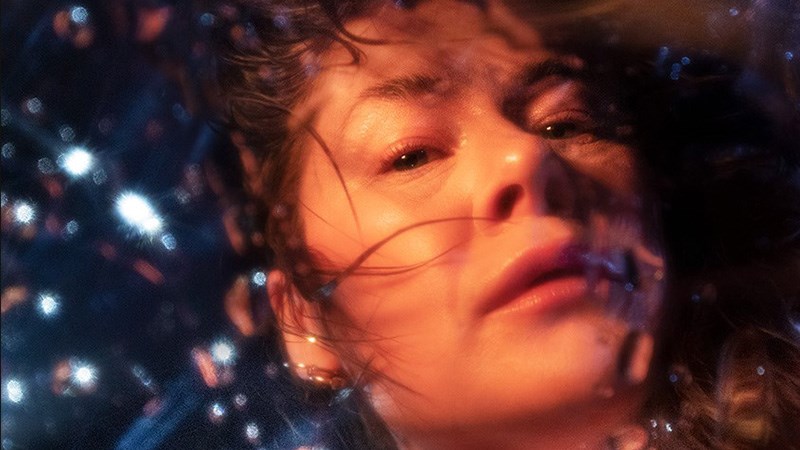 En kvinnas ansikte mot en abstrakt bakgrund som ser ut att vara under vatten. Hon tittar in i kameran med en allvarlig, lite vemodig, min.)