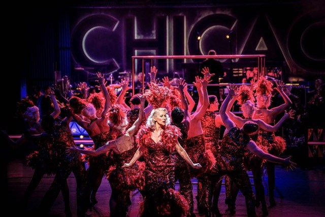 Shownummer från föreställningen Chicago på Malmö Opera. I förgrunden en kvinna i röd klänning och bakom henne en dansande ensemble. Scenen är ljussatt med rött ljus.