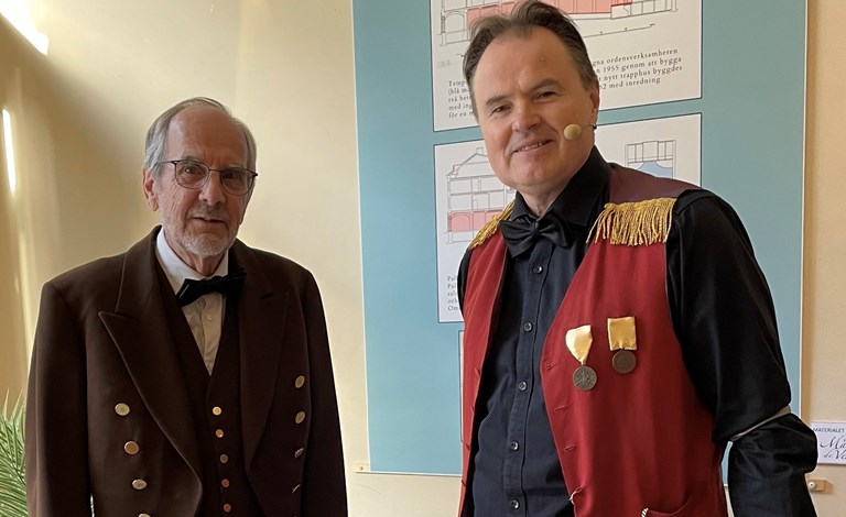 En bild föreställande två män. Den ena bär en brun uniform med guldknappar, den andra bär en röd väst med guldfransar på axlarna och medaljer på västbröstet.