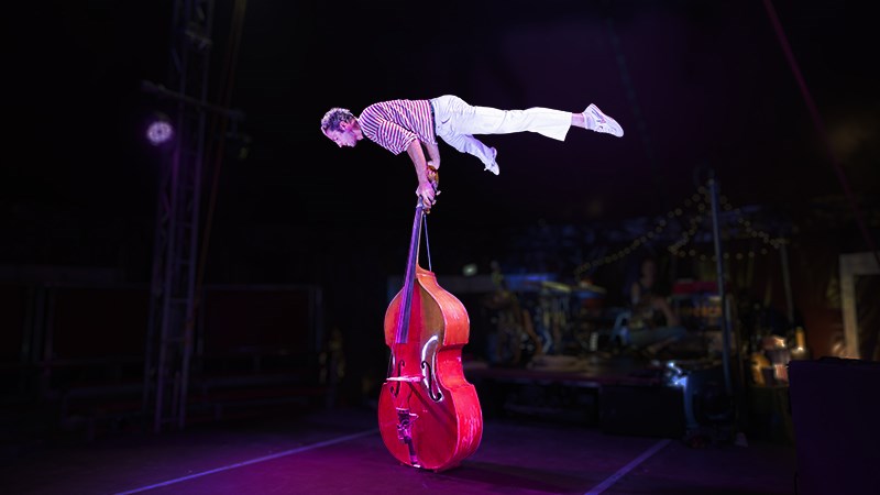 En scen där en cirkusartist balanserar vågrätt på en stående kontrabas.)