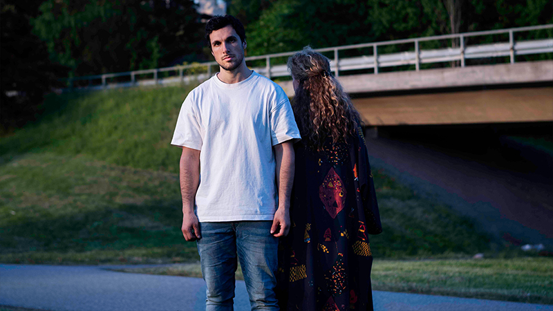 De två medverkande i en urban miljö. Mannen tittar in i kameran, kvinnan står bredvid honom med ryggen mot betraktaren.)