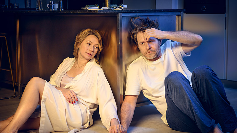 Frida Hallgren och Henrik Norlén i karaktär, sitter på golvet och håller varandra i handen.)