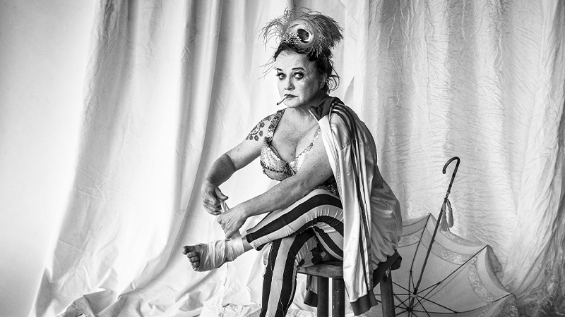 Foto på skådespelaren i karaktär. Hon är uppklädd och har håret uppsatt, och en cigarrett i mungipan, och sitter och lindar sin vänsterfot.)