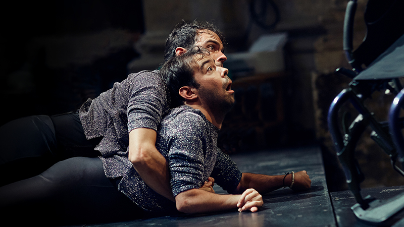 Två män i likadana svarta byxor och glittriga tröjor. Den ena ligger på mage på golvet, och den andra håller sina armar runt honom. Till höger en rullstol.)