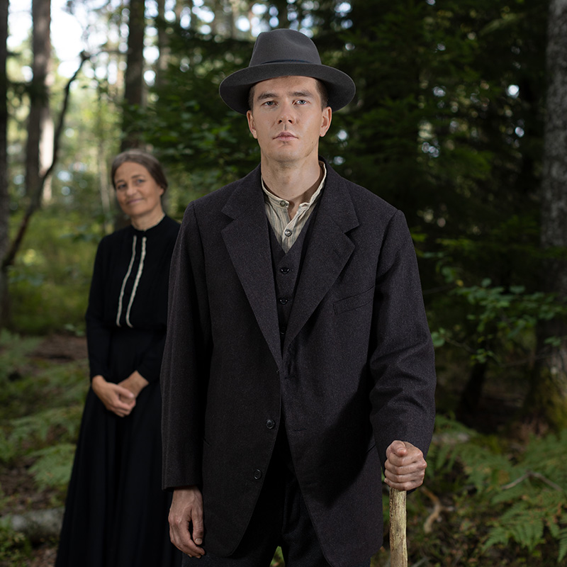 Personer i karaktär står i en skog: i förgrunden Aki Pirkola med en träkäpp, och i bakgrunden Mette Marqvardsen.