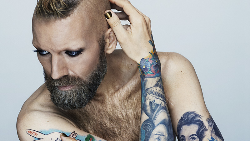 Rickard Söderberg lutar sig mot ett bord. Vi ser hans nakna överkropp, och armarna med tatuerade "sleeves" med ansikten i blått med färgglada detaljer.)