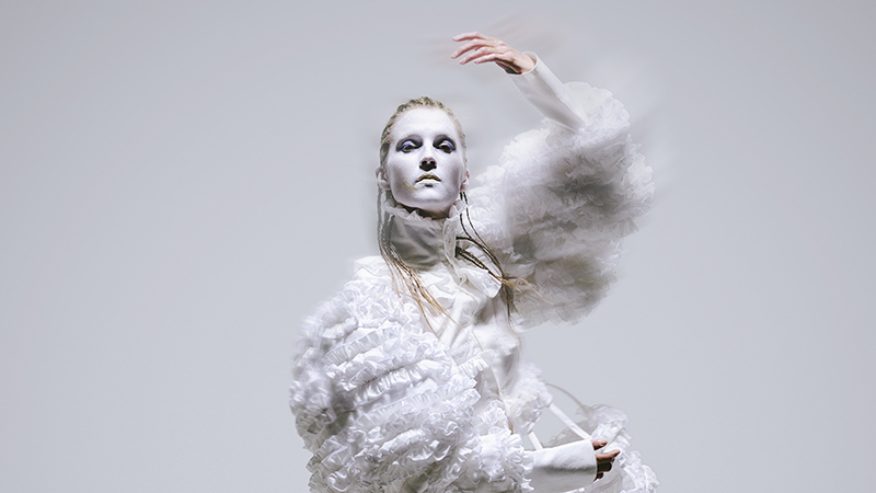 Frauke fångad mitt i en dansrörelse. Hennes klädsel har ränder av krusiduller och hennes ansikte är vitsminkat. )