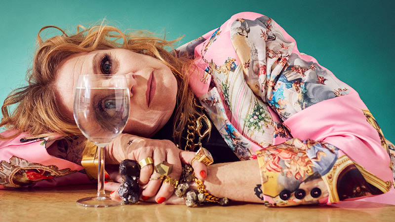 En kvinna i färgglad blus ligger med huvudet på bordet och tittar genom ett fullt vinglas.)